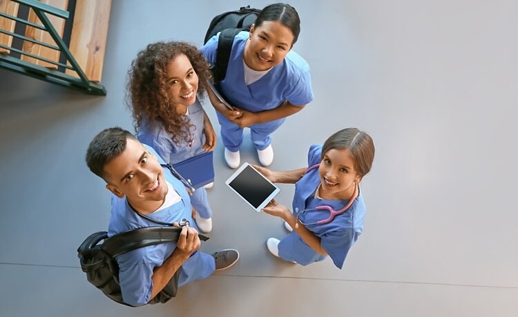 Kleine Gruppe von Studenten im Pflegebereich stehen zusammen und schauen lächelnd nach oben in die Linse für das Gruppenbild. 