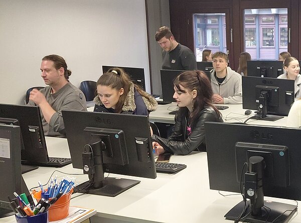 In einem modernen Computerraum der Universitätsbibliothek Würzburg sitzen mehrere angehende Erzieher der Euro Akademie Tauberbischofsheim an weißen Tischen mit schwarzen Desktop-Computern. Die Personen sind in verschiedenen Sitzpositionen zu sehen, einige arbeiten konzentriert an den Bildschirmen, andere unterhalten sich. Im Hintergrund sind große Fenster mit Blick auf eine historische Fassade zu erkennen. Auf einem Tisch im Vordergrund steht ein orangefarbener Behälter mit zahlreichen blauen und schwarzen Stiften. Die Atmosphäre wirkt ruhig und lernorientiert, typisch für eine Exkursion in einer Bibliothek.