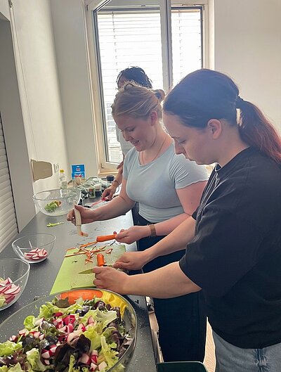 Drei Auszubildende stehen nebeneinander an einer Küchenarbeitsplatte und schneiden Gemüse. Im Vordergrund liegen auf einem Schneidebrett Karotten und ein Sparschäler, daneben eine große Glasschüssel mit gemischtem Salat. Im Hintergrund fällt Tageslicht durch ein Fenster mit Jalousien.