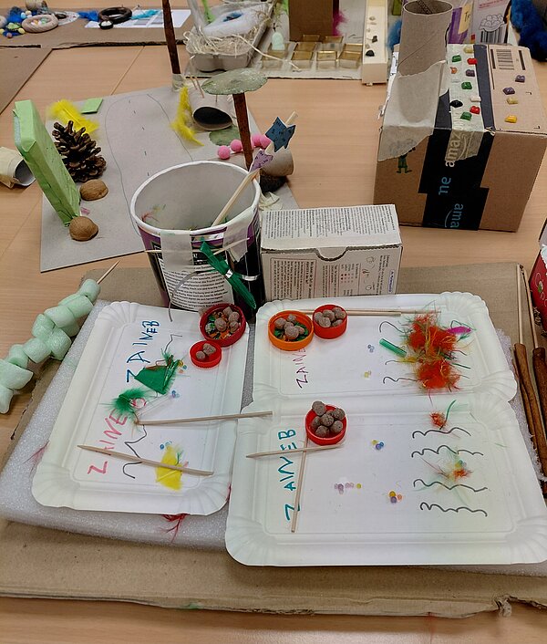 Auf zwei weißen Papptellern liegen kleine Bastelarrangements aus bunten Federn, Zahnstochern, Kügelchen und gefüllten roten Schraubdeckeln, daneben stehen Becher und Kartons auf einer großen Pappunterlage inmitten anderer Projekte.