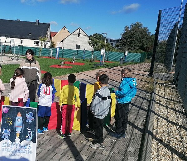 Kinder stehen hinter einem bunten Spielgerät im Freien, daneben ein Schild ‚Neptuns Sternenreise – Station 6' mit Astronauten-Illustration; im Hintergrund Spielplatz und Wohnhäuser bei Sonnenschein.