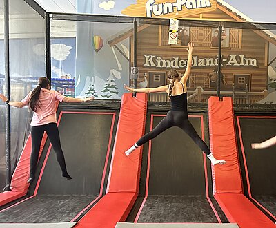 Zwei junge Frauen springen hoch auf einem Trampolin mit schrägen schwarz-roten Streifen. Das Foto ist von hinten aufgenommen.