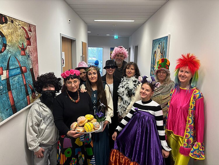 Gruppe von verkleideten Personen in einem hellen Flur, einige mit Perücken, Hüten und bunten Kostümen; vorne hält eine Person ein Tablett mit Berlinern/Krapfen in die Kamera.