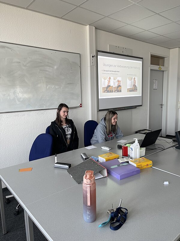 Zwei weibliche Auszubildende sitzen in einem Klassenzimmer an einem Tisch. Eine Person trägt einen schwarzen Kapuzenpullover, die andere ein graues Sweatshirt und benutzt einen Laptop. Auf dem Tisch liegen verschiedene Gegenstände wie eine pinkfarbene Wasserflasche, Schlüssel, Ordner und Putzmittel. Auf einer Leinwand wird eine Präsentation mit dem Titel „Übungen zur Verbesserung der Haltung“ gezeigt, die Illustrationen von Übungen im Sitzen enthält. Der Raum ist mit einer Tafel und Bürostühlen ausgestattet.