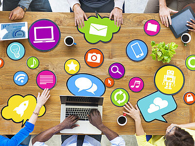 Mehrere Personen sitzen um einen Tisch herum, auf dem ein Laptop und verschiedene farbige Symbole für Technologie und Kommunikation liegen, wie z.B. ein Computer, ein Briefumschlag, eine Kamera, ein Smartphone und Wolken. Die Personen scheinen an einer Besprechung teilzunehmen und interagieren mit den Symbolen.