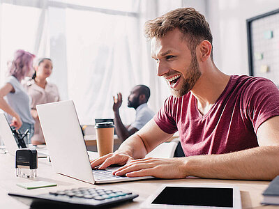 Ein junger Mann in rotem T-Shirt sitzt an einem Schreibtisch mit Notebook. Dahinter sind zwei Frauen und ein weiterer Mann, die sich unterhalten.