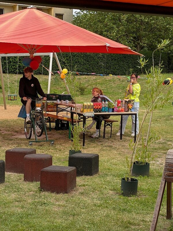 Unter einem Sonnenschirm ist eine kleine Station mit Fahrrad-Mixer, Getränken und Zutaten für Smoothies aufgebaut.