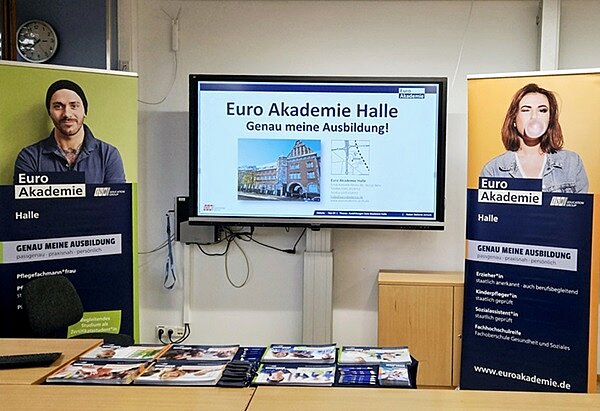 Messestand der Euro Akademie Halle mit großem Bildschirm in der Mitte, zwei großen Rollups links und rechts und einem Tisch mit Broschüren.