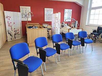 In einem Klassenraum stehen vorne fünf blaue Stühle mit Armlehnen; im Hintergrund sind zwei Übungs-Krankenbetten sowie ein Rollstuhl zu sehen.