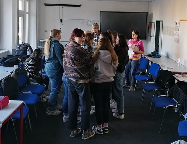 Sozialassistent*innen vertiefen Wissen über Cybermobbing Eine Gruppe von Schülern und Schülerinnen steht eng beisammen in einem Klassenzimmer, während eine Lehrkraft im Hintergrund Anweisungen gibt. Die Szene zeigt eine interaktive Übung im Rahmen einer pädagogischen Nachbereitung zum Thema Cybermobbing.