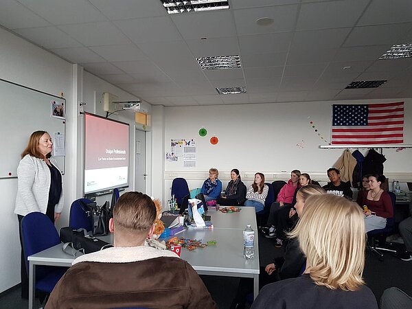Career Opportunities an der Euro Akademie - Einblicke in die Berufswelt Eine Frau hält einen Vortrag mit einer Präsentation mit dem Titel „Unique Professionals“. Die Klasse sitzt an Gruppentischen, an der Wand hängt eine große US-Flagge. Auf den Tischen stehen Snacks und Getränke.