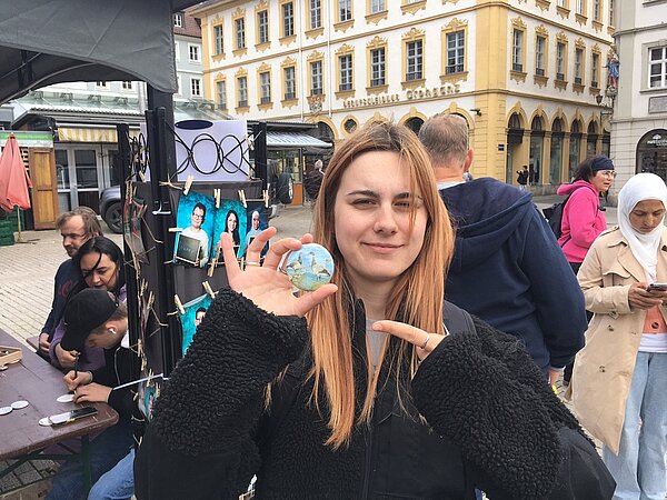 Protesttag zur Gleichstellung von Menschen mit Behinderung: Buttons für Inklusion Eine Schülerin hält einen Button, auf dem zwei Nilgänse abgebildet sind, in die Kamera und zeigt mit ihrem Finger auf das Motiv. Dahinter sind Passant*innen erkennbar und das Geschäft "Berufsbekleidung Grohganz".