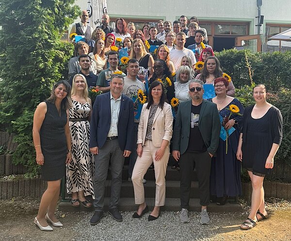 Pflege mit Herz, Verstand und Haltung – Ein bewegender Abschluss an der Euro Akademie Jena Eine große Gruppe von Absolvent*innen und Gästen posiert lächelnd auf einer Treppe vor einem Gebäude. Viele halten Sonnenblumen und Urkunden in den Händen. Das Bild zeigt die Abschlussfeier der Pflegefachkräfte an der Euro Akademie Jena.