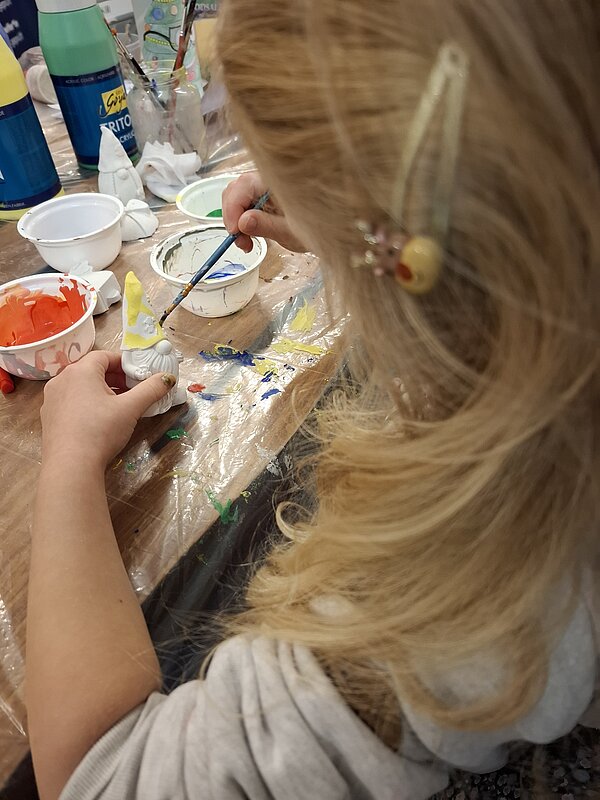 Ein blondes Kind sitzt an einem Tisch und bemalt konzentriert eine kleine weiße Weihnachtsfigur mit gelber Acrylfarbe, umgeben von bunten Farbbechern und Pinseln.