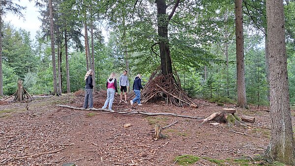 Vier Jugendliche in einem Waldstück beim gemeinsamen Bau einer einfachen Hütte aus Ästen und Zweigen rund um einen Baum.