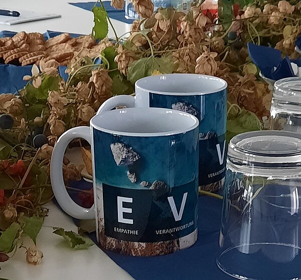 Zwei Tassen stehen auf dem Tisch, davor zwei Wassergläser, ein Teller Kekse und ein herbstlicher Ast mit Blättern.