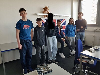 Sechs Jugendliche stehen in einem Klassenraum vor einer aufgehängten britischen Flagge; eine Person in der Mitte ist unkenntlich gemacht.