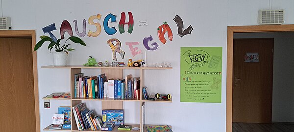 An einer weißen Wand steht in bunten Buchstaben das Wort "Tauschregal", darunter befindet sich ein Regal mit verschiedenen Gegenständen, darunter Kuscheltiere, Spiele, Bücher und eine Pflanze. Daneben hängt ein Poster, das die Tauschregeln erklärt.