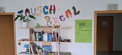 An einer weißen Wand steht in bunten Buchstaben das Wort "Tauschregal", darunter befindet sich ein Regal mit verschiedenen Gegenständen, darunter Kuscheltiere, Spiele, Bücher und eine Pflanze. Daneben hängt ein Poster, das die Tauschregeln erklärt.