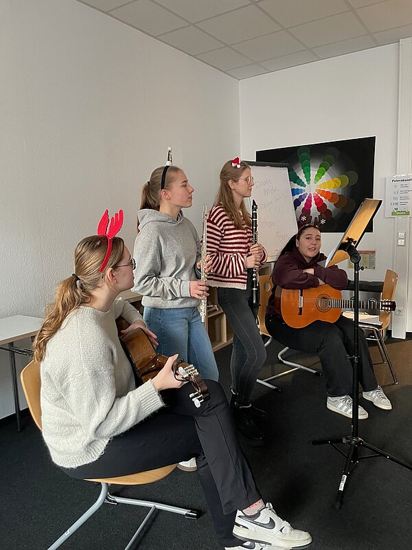 Eine kleine Gruppe Studierender mit Klarinetten, Gitarre und Ukulele sitzt in einem Klassenraum und musiziert gemeinsam mit weihnachtlichen Accessoires wie roten Haarreifen.