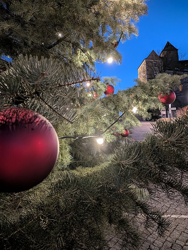 Nahaufnahme von Tannenzweigen mit roten Christbaumkugeln und Lichtern, im Hintergrund eine Burg und gepflasterter Platz bei Abendlicht.