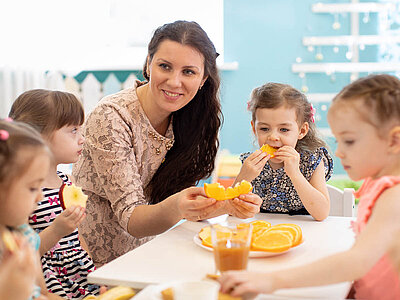 Eine Kindergruppe mit einer Erzieherin teilt Orangenscheiben an einem Tisch, während im Hintergrund eine Glas Orangensaft steht.