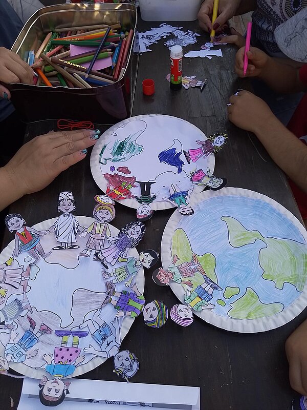 Ausgemalte und ausgeschnittene Papierfiguren werden auf Papptellern mit Weltkarten aufgeklebt, sodass eine Menschenkette um die Erde entsteht.