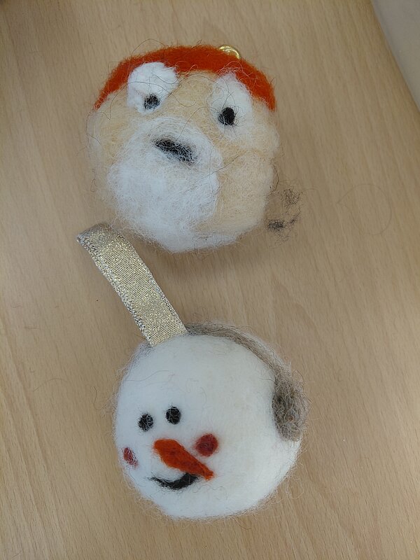 Zwei runde, aus Filzwolle gefertigte Weihnachtsanhänger auf einem Holztisch: oben ein Gesicht mit roter Mütze und weißem Bart, unten ein Schneemannkopf mit orangefarbener Nase, roten Wangen und goldenem Aufhängeband.