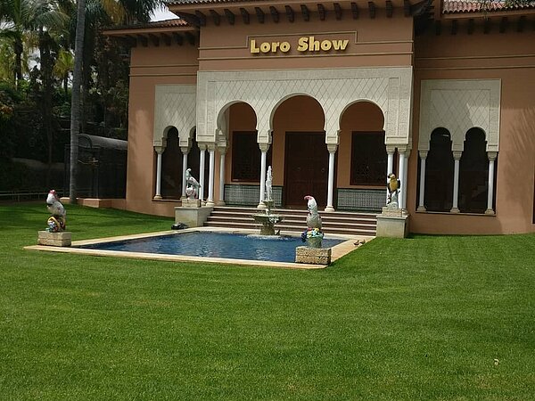 Gebäude mit weißen Säulen und großem Eingangsbereich, auf dem in goldener Schrift "Loro Show" geschrieben steht. Davor befindet sich ein Pool, an dessen Ecken Skulpturen von verschiedenen Papageienvögeln stehen. Der Pool ist von einer grünen Grasfläche umgeben.