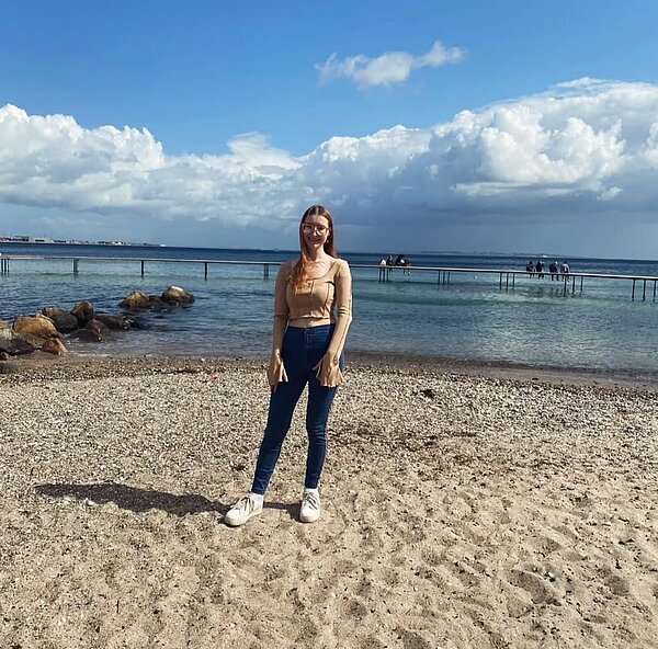 Erfahrungsbericht Erasmus – Hygge von Janice Weßel Eine junge Frau mit langen, hellbraunen Haaren steht lächelnd auf einem Kiesstrand mit Blick auf das Meer. Sie trägt eine beige Bluse, blaue Jeans und weiße Sneakers. Hinter ihr führt ein langer Steg ins Wasser, auf dem mehrere Menschen sitzen. Der Himmel ist blau mit einigen weißen Wolken.