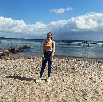 Eine junge Frau mit langen, hellbraunen Haaren steht lächelnd auf einem Kiesstrand mit Blick auf das Meer. Sie trägt eine beige Bluse, blaue Jeans und weiße Sneakers. Hinter ihr führt ein langer Steg ins Wasser, auf dem mehrere Menschen sitzen. Der Himmel ist blau mit einigen weißen Wolken.