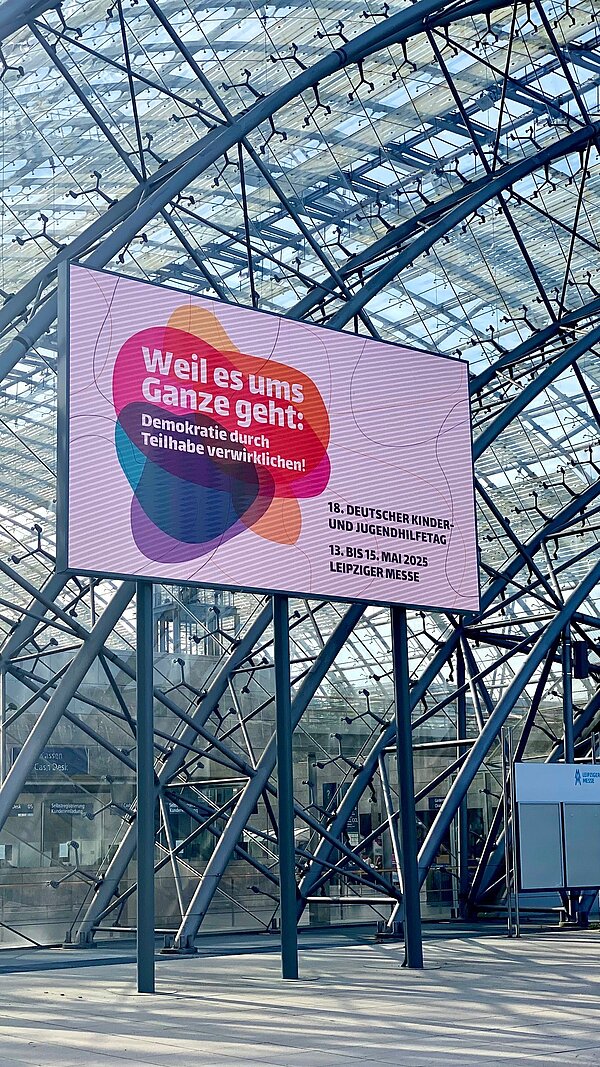Deutscher Kinder- und Jugendhilfetag 2025: „Heute laut, morgen stark“ Die Messehalle mit Glaskuppel, darin eine Reklametafel, auf der für die Messe geworben wird: "Weil es ums Ganze geht: Demokratie durch Teilhabe verwirklichen! 18. Deutscher Kinder- und Jugendhilfetag · 13. bis 15. Mai 2025 · Leipziger Messe"