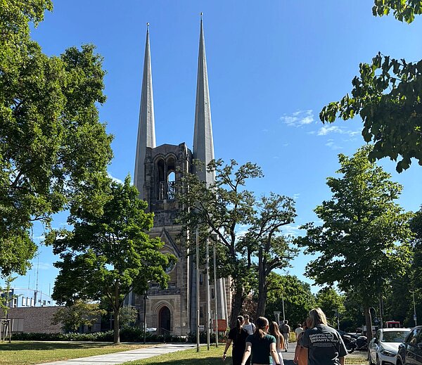 Gruppe von Menschen spaziert auf einem Gehweg in Richtung einer Kirche mit zwei markanten, hohen Kirchtürmen an einem sonnigen Tag.
