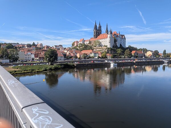 Panorama einer historischen Stadt am Fluss mit einer großen Burg- und Kirchenanlage auf einem Hügel. Die Gebäude und der blaue Himmel spiegeln sich im ruhigen Wasser.