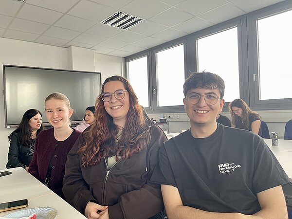 Absolventinnen und Absolventen der Euro Akademie sitzen gut gelaunt in einem hellen Raum an weißen Tischen. Im Vordergrund sind drei Personen zu sehen, die nebeneinander sitzen. Im Hintergrund befinden sich weitere Personen, die an einem Tisch arbeiten oder miteinander sprechen. Große Fenster lassen Tageslicht herein, und an der Wand hängt ein großer, ausgeschalteter Bildschirm. Die Decke ist mit weißen Paneelen und Leuchtstofflampen ausgestattet.