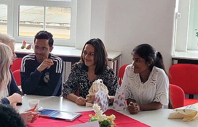 Drei Personen sitzen an einem Tisch in einem Seminarraum und unterhalten sich mit zwei weiteren Personen, die nur zur Hälfte zu sehen sind. Auf dem Tisch liegen kleine Geschenktüten und ein Heft. Im Hintergrund sind Fenster und ein Teil eines Gebäudes zu sehen.