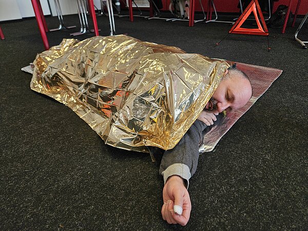 Nahaufnahme eines Teilnehmers, der in eine goldene Rettungsdecke eingewickelt seitlich auf einer Matte liegt; Hand mit Pflaster sichtbar, Warndreieck hinten.