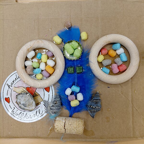 Nahaufnahme einer Collage auf Karton: Zwei Holzringe sind mit farbigen Kügelchen gefüllt, dazwischen sitzt eine blaue Feder mit weiteren Kügelchen, Muscheln und ein Korken, sodass eine symmetrische, fast maskenartige Form entsteht.