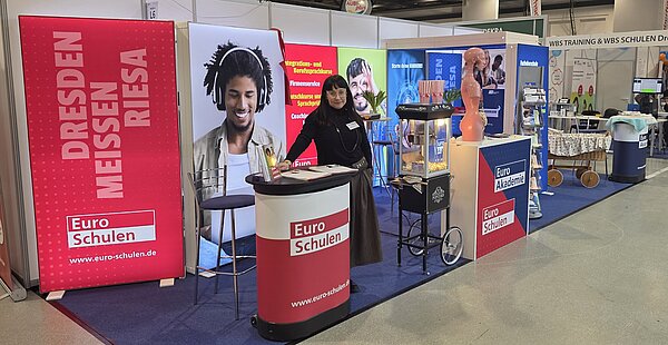Eine Frau in dunklem Rock am Messestand der Euro-Schulen und Euro Akademie; dort stehen unter anderem ein Popcornwagen und eine Anatomiepuppe