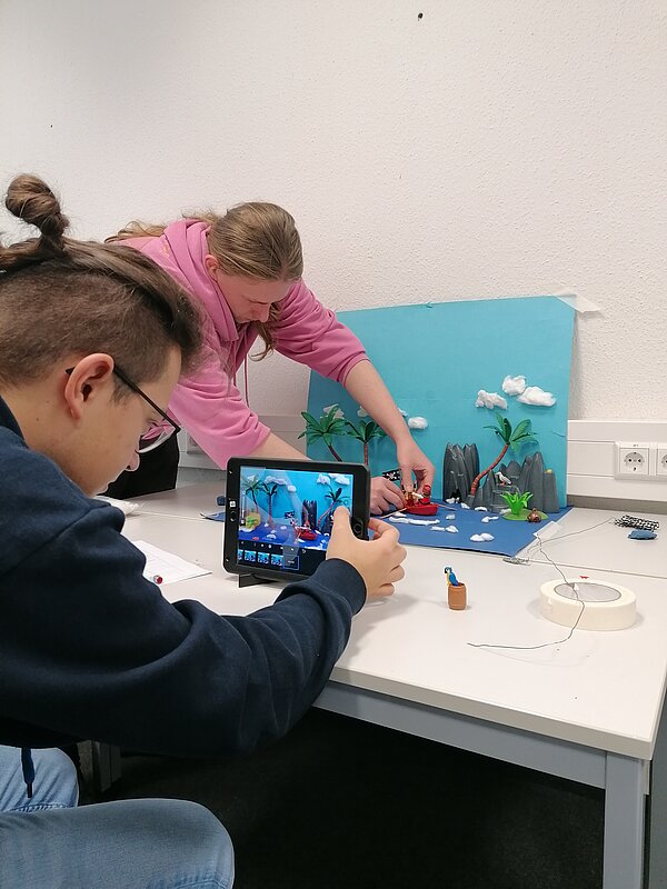 MeKoSax-Projekt „Trickfilmproduktion“ Eine Schülerin filmt eine aus Playmobil gebaute Küste mit Palmen und Felsen, welche auf einem hellbraunen Stück Papier stehen und vor einem hellblauen Hintergrund mit Wolken.