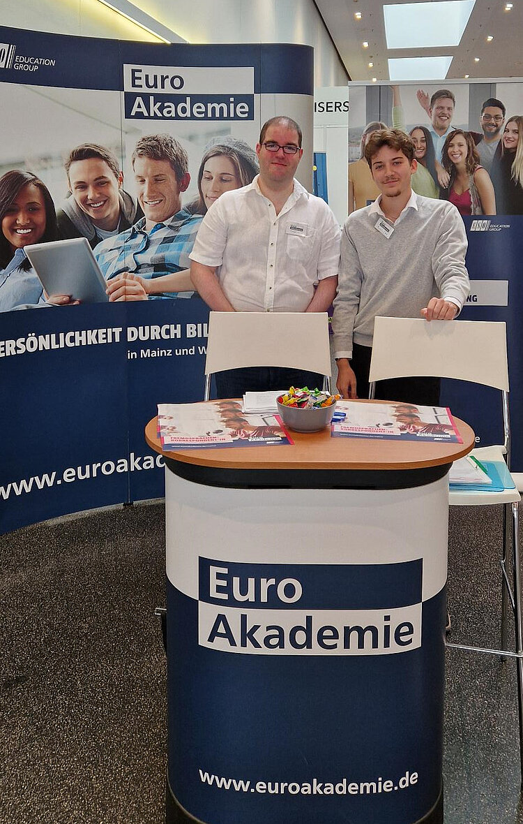 Zwei Männer in hellen Hemden stehen hinter einem runden Tisch mit der Aufschrift 'Euro Akademie' und der Website www.euroakademie.de. Auf dem Tisch liegen Informationsmaterialien und ein Tablett mit Snacks. Im Hintergrund sind Roll-ups mit Bildern von Studierenden und dem Slogan 'Persönlichkeit durch Bildung' zu sehen.