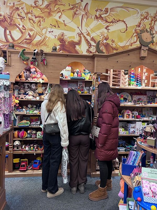 Drei Personen stehen in einem Spielzeugladen und betrachten verschiedene Spielzeuge, die auf Regalen und Tischen ausgestellt sind.