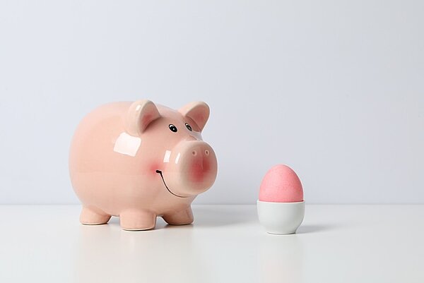 Ein rosa Sparschwein steht auf einer hellen Oberfläche neben einem rosa Ei, das in einem weißen Eierbecher steckt. Der Hintergrund ist schlicht und weiß gehalten.