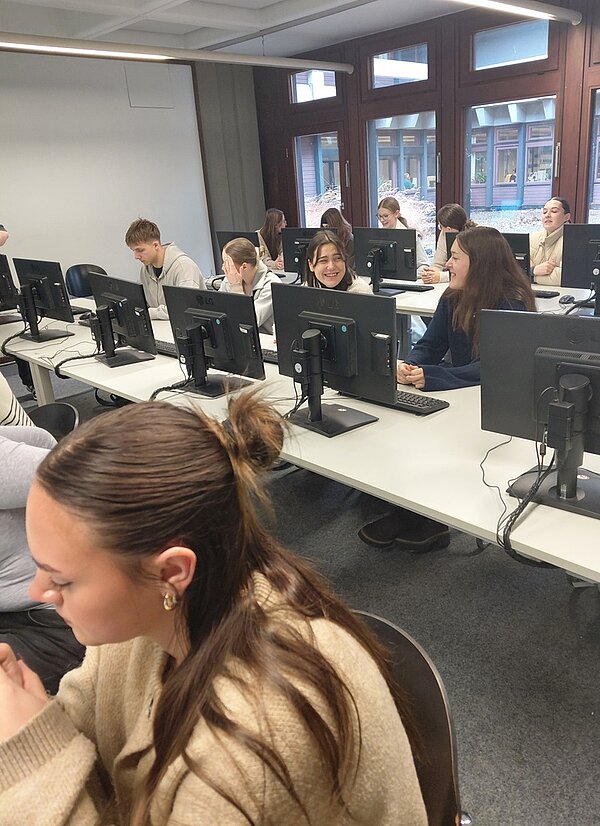 In einem Computerraum der Universitätsbibliothek Würzburg sitzen mehrere angehende Erzieher der Euro Akademie Tauberbischofsheim an langen Tischen mit mehreren Desktop-Computern. Die Personen sind in Reihen angeordnet und konzentrieren sich auf die Bildschirme vor ihnen. Der Raum ist hell beleuchtet, mit großen Fenstern an der rechten Seite, durch die man die Fassade eines benachbarten Gebäudes sehen kann. Die Decke ist mit weißen Paneelen verkleidet, und die Wände sind hell gestrichen. Die Tische sind weiß, die Stühle dunkel und schlicht gestaltet. Die Atmosphäre wirkt ruhig und lernorientiert.