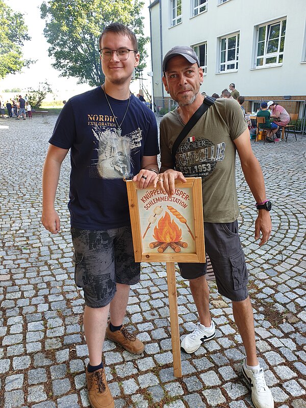 Ein erlebnisreicher Vormittag voller Spiel, Kreativität und Gemeinschaft Zwei Männer stehen vor einem Schild mit der Aufschrift „Knüppelkuchen-Station“, das ein Lagerfeuer und Marshmallows zeigt.