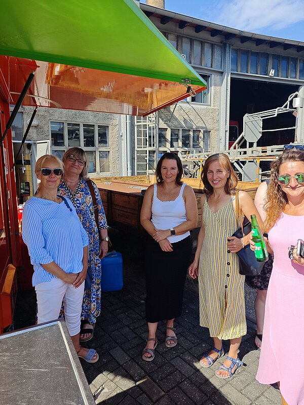 Fünf Frauen in sommerlicher Kleidung unter einem grünen Dach und orangefarbener Unterseite. Sie lächeln in die Kamera und wirken entspannt. Eine der Frauen hält eine Flasche in der Hand. Im Hintergrund sind weitere Gebäude und technische Konstruktionen zu sehen.