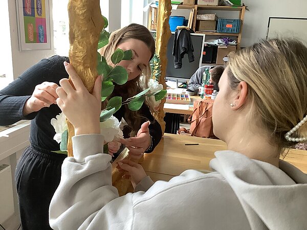 Zwei Schülerinnen befestigen ein Blumenbouquet an der Ecke des Bilderrahmens.