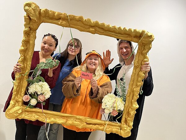 Vier Studierende schauen lächelnd in ihren Faschingskostümen durch einen übergroßen selbst gestalteten Bilderrahmen und posieren für ein Foto.