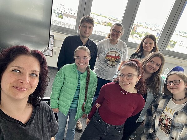 Eine Gruppe von Absolventinnen und Absolventen posiert fröhlich für ein Selfie. Im Hintergrund sieht man aus dem Fenster die Stadt Aschaffenburg.