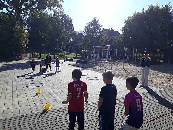 Kinder stehen an für den Parcours, bei dem Pylonen aufgebaut sind und Hula Hoop Reifen auf dem Boden liegen.